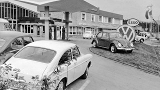 Schröder Team - Alles begann mit einer Tankstelle Eine schwarzweiß Außenaufnahme der Tankstelle und Werkstattgebäude. Mehrere Autos aus der Zeit stehen verteilt auf dem Hof.