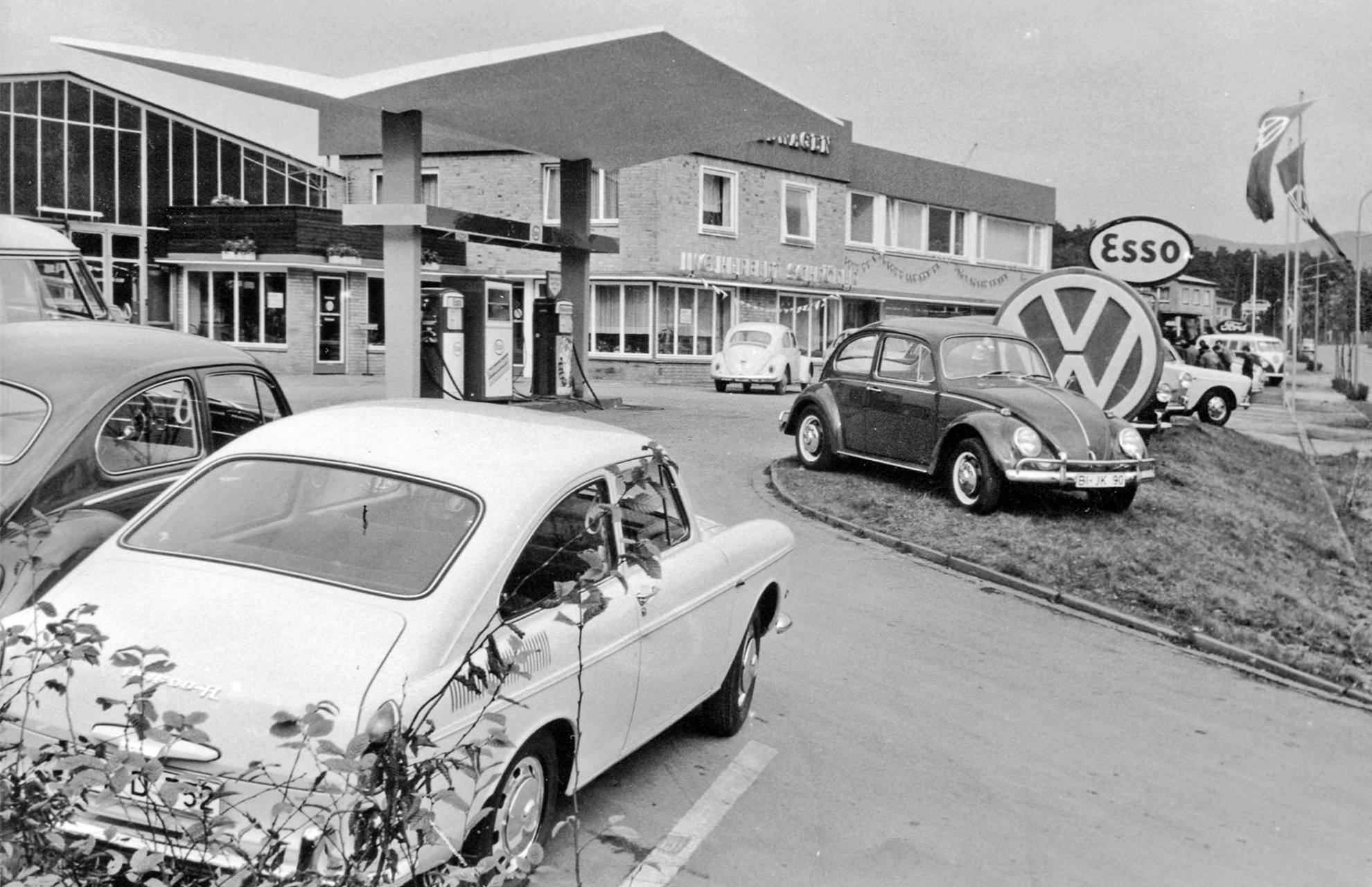 Schröder Team - Alles begann mit einer Tankstelle Eine schwarzweiß Außenaufnahme der Tankstelle und Werkstattgebäude. Mehrere Autos aus der Zeit stehen verteilt auf dem Hof.